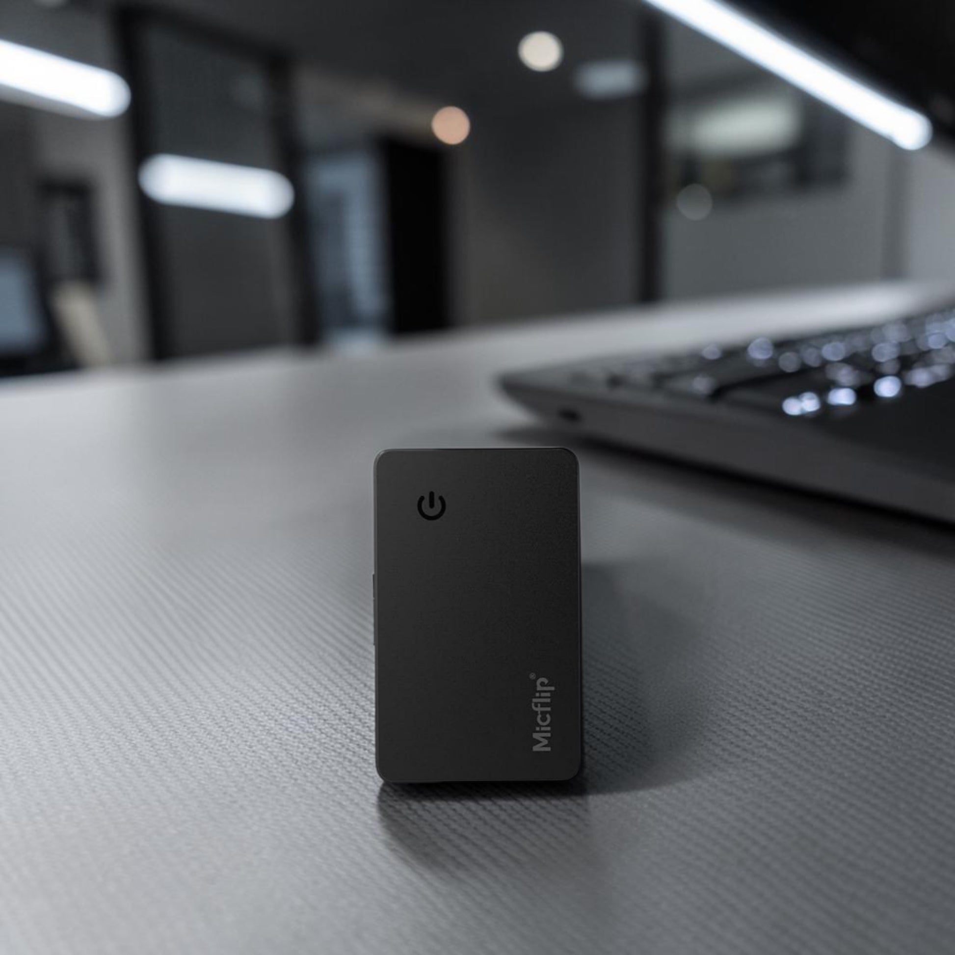 Micflip Ultra-Thin Wallet Card Finder in black, slim card-shaped device with Micflip branding, standing on a desk with a blurred laptop and office lights in the background.